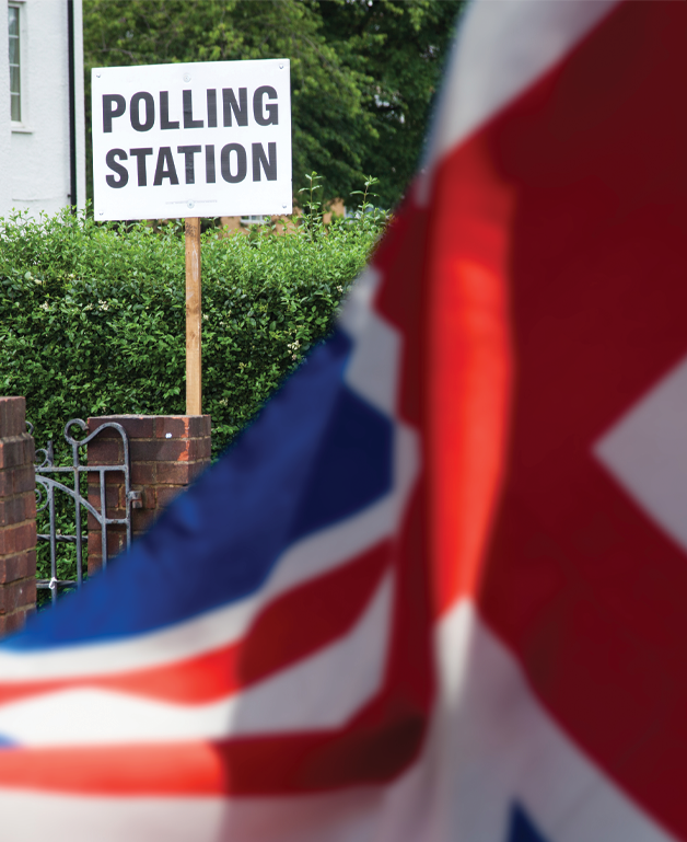 A polling station sign is in front of a white building. To the righthand side a Union Jack flag is blowing in the breeze.