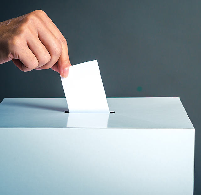 someone's hand is reaching over a white box posting a voting card against a dark grey backdrop