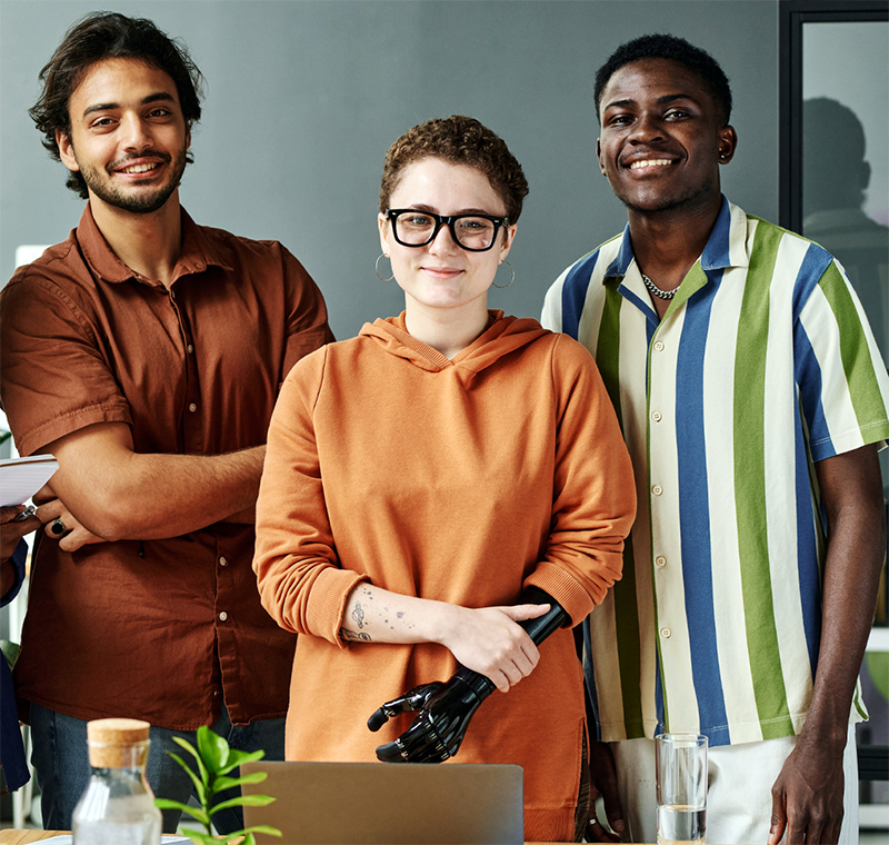 Three work colleagues are stood together in an office space looking happy. In between a Black and Asian smiley man, is a young white female who is wearing glasses and has a black prosthetic arm, who is in front of a laptop.