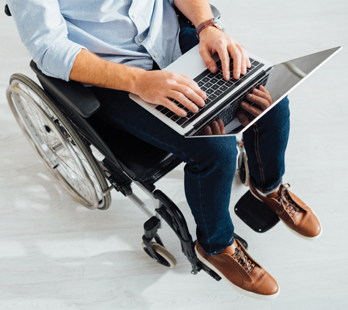 A male wheelchair user, in office attire, has a laptop on his knee that he is typing into.