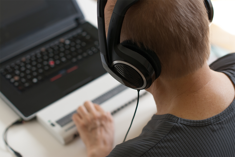 Taken from behind, there is a man with short brown hair, who is wearing a black t-shirt and headphones, is sitting at a desk typing on a Braille keyboard attached to a laptop.